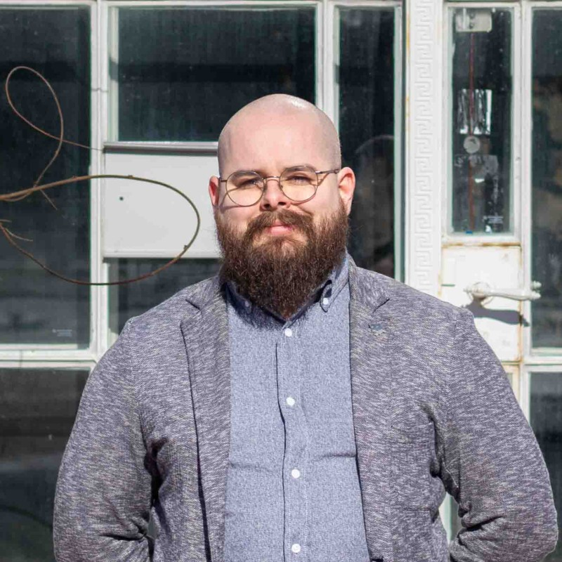 A man with beard and glasses wearing casual suit and posing with little smile for taking picture.